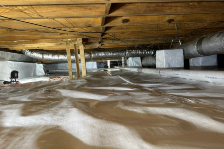 A clean, encapsulated crawl space with a white reinforced vapor barrier covering the floor and walls. Exposed wooden floor joists and insulation ducts run across the ceiling. A dehumidifier and drainage system components are visible, ensuring moisture control and improved air quality.